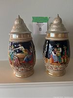 Front views of two decorative ceramic beer steins with pewter lids showing colorful traditional Bavarian scenes