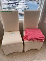 Two cream accent chairs side-by-side, one with pink throw blanket on seat, near window with marina view