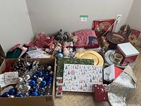 Photo of assorted Christmas decorations including ornaments, charger plates, decorative boxes, fabric napkins, and wrapping papers arranged on floor.