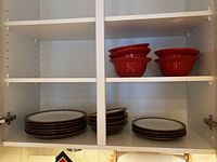 Cabinet shelf with stacked Denby plates, white with brown rims. Four white bowls with brown rims stacked and four detached red ceramic bowls stacked.