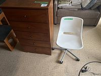 Wooden dresser with four drawers beside a small white office chair on wheels, placed on carpeted floor.