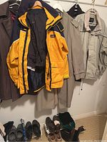 Several men's coats hanging on a rack including a yellow and blue Gill jacket and various other coats; shoes and boots visible on floor, showing condition and variety.