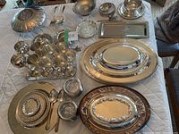Photo displaying full assortment of silver-tone serving trays, bowls, cups, flatware, and decorative pieces laid out on quilted surface.