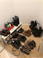 A variety of men's and women's shoes, including casual, hiking, and rubber boots arranged near and on a white two-tier shoe rack. A red tray on the rack holds shoe care products.