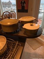 Photo showing three items on a round table: the avocado green electric pot with lid, the Pyrex casserole with patterned lid on a metal stand, and the Corning Ware casserole pot with white lid.