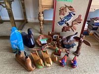 Wide view of wood carvings and framed needlepoint picture arranged on floor, showing all items in the lot