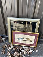 Two framed artworks outdoors against metal wall, fallen leaves in foreground. Includes Egyptian theme framed in glass and wild fowl scene framed in gold-tone faux bamboo.