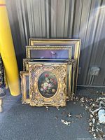 Grouped view of five framed floral artworks with ornate gold frames leaning against wall outdoors.