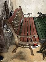 Side angled view of bench with red wooden slats and wrought iron frame showing lattice backrest and scrollwork on armrest and legs.