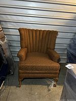 Front view of a vintage brown upholstered chair with scalloped back and curved armrests, wooden carved legs heavily scratched.