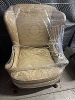 Beige upholstered wingback chair wrapped in plastic, showing overall shape and fabric condition.