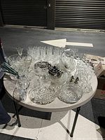 Table top display of assorted clear glassware including bowls, glasses, salt and pepper shakers, crystal vases, and silver-plated teapot on glass tray