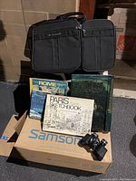 Photo showing Samsonite black soft side carry-on bag atop a Samsonite box with an assortment of travel and cookbook books and Tasko binoculars in front, placed on concrete steps.