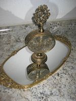 Ormolu mirrored vanity tray with ornate floral designs on handles and edges, holding a decorative glass perfume bottle with floral gilt pattern and floral top.
