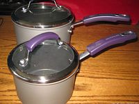 Two Rachael Ray saucepans with glass lids and purple handles on wooden surface.