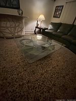 Square glass coffee table with sculptural abstract base under the glass, shown from a corner angle near a brown leather sofa and side table with lamp.