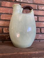 Light green beige pottery jug with an oval body, narrow neck, a spout and handle wrapped in dark woven material.