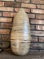 Full view of the tall pottery vase placed against a brick wall background showing its full shape and color patterning.