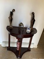 Photo showing a wooden half-moon entrance table against a white wall, decorated with two abstract bronze-tone human figurines, a bronze-tone deer figurine, a small white ceramic jar with a blue rim on top, and a large ceramic vase with a geometric pattern on the lower shelf.