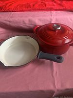 View of the red Dutch oven with lid and frying pan on a pink surface showing color and finish.