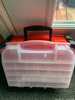 Photo showing clear plastic compartmentalized storage box with black handle and red sides in the background.