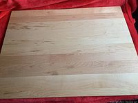 Photo showing large rectangular wooden cutting board with natural wood grain on a red fabric background.