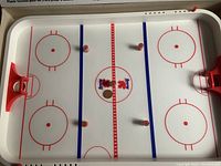 Top-down view of the white plastic hockey rink table game with red goals and rink markings showing the playing surface and four pegs.