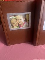 Two brown leatherette photo albums standing upright showing the front photo insert window with a photo of two young girls, album covers stitched around edges.
