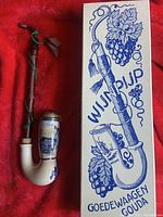 Ceramic wine pipe with decorative blue scenes and wooden stem, displayed beside original box with grapevine graphics.