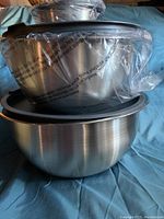 Three stainless steel bowls stacked, each with a dark plastic lid, partially covered in plastic bag.