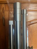 Photo showing three metal double drapery rods with cylindrical ends in sealed packaging, silver finish.