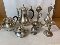Full lot photo showing all seven pewter items on white background: coffee pot, teapot, sugar bowl, creamer, lidded vessel, candlestick holder, and pitcher.