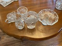 Photo showing six crystal and glass items on a wooden table including vases, a bowl, and a creamer-style pitcher with cut patterns.