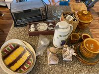 Overview of lot including small vintage toaster oven, ceramic serving pieces in various colors, MCM ceramic bookends shaped as figures, glassware, wicker basket with small items, and ceramic coffee pot