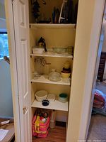 Shelf with stacked glass bowls, Corningware casserole dish, assorted kitchen storage containers and colorful plates or lids.