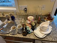 Overview of lot showing glasses, mugs, ceramic pitcher with wicker handle, silver tray, and white ceramic serving dishes.