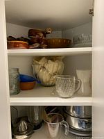 Photo showing various glassware, soup bowls, small colored bowls, pitcher, and metal cookware within a cabinet.