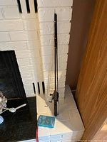Full view of Shimano fishing rod standing against white brick wall with a small box of lures on floor in front