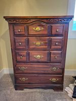 Full frontal photo of solid wood tallboy chest showing carved top edge, five drawers with brass handles and knobs, and wooden paneling.