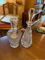 Two crystal decanters displayed on a wooden surface with background furniture visible.