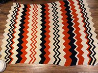Full view of the vintage crocheted blanket laid flat on wooden floor, showing multicoloured zigzag pattern in cream, orange, rust, and black.