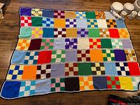 Full view of the crocheted blanket laid flat on wooden floor, showing rows of various colored square patches in checker and solid patterns.