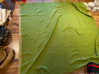 Full view of the green wool blanket spread on the floor showing its size, color, and beige border trim.