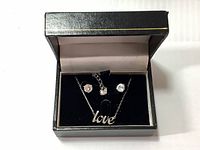 Open black jewelry box displaying pair of Swarovski solitaire earrings and 'love' pendant necklace against black velvet