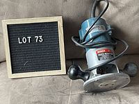 Bosch 1602 electric router placed beside a black sign with white letters reading 'LOT 73'. Router is blue and silver with black handles and has a power cord coiled on top.