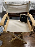 Front view of the director's chair showing creamy white canvas and light wood frame, with semitransparent backrest and armrests. Chair is on wooden floor and includes a small blackboard with 'Lot 63'.