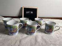 Six Capilano mugs displayed on a towel with a sign 'LOT 50' in background. Mugs feature a floral and window scene design in mostly blue, green, and red hues.