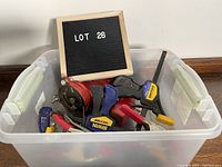 Photo showing a plastic bin containing various clamps and one red bench vise with a sign 'LOT 26'