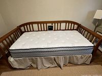 Wooden daybed against a light-colored wall, showing the mattress with a beige bed skirt and wooden slatted back and sides.
