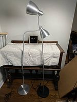 Two metal floor lamps, one with silver base, one with black base, standing side by side on hardwood floor against white wall and covered furniture behind.